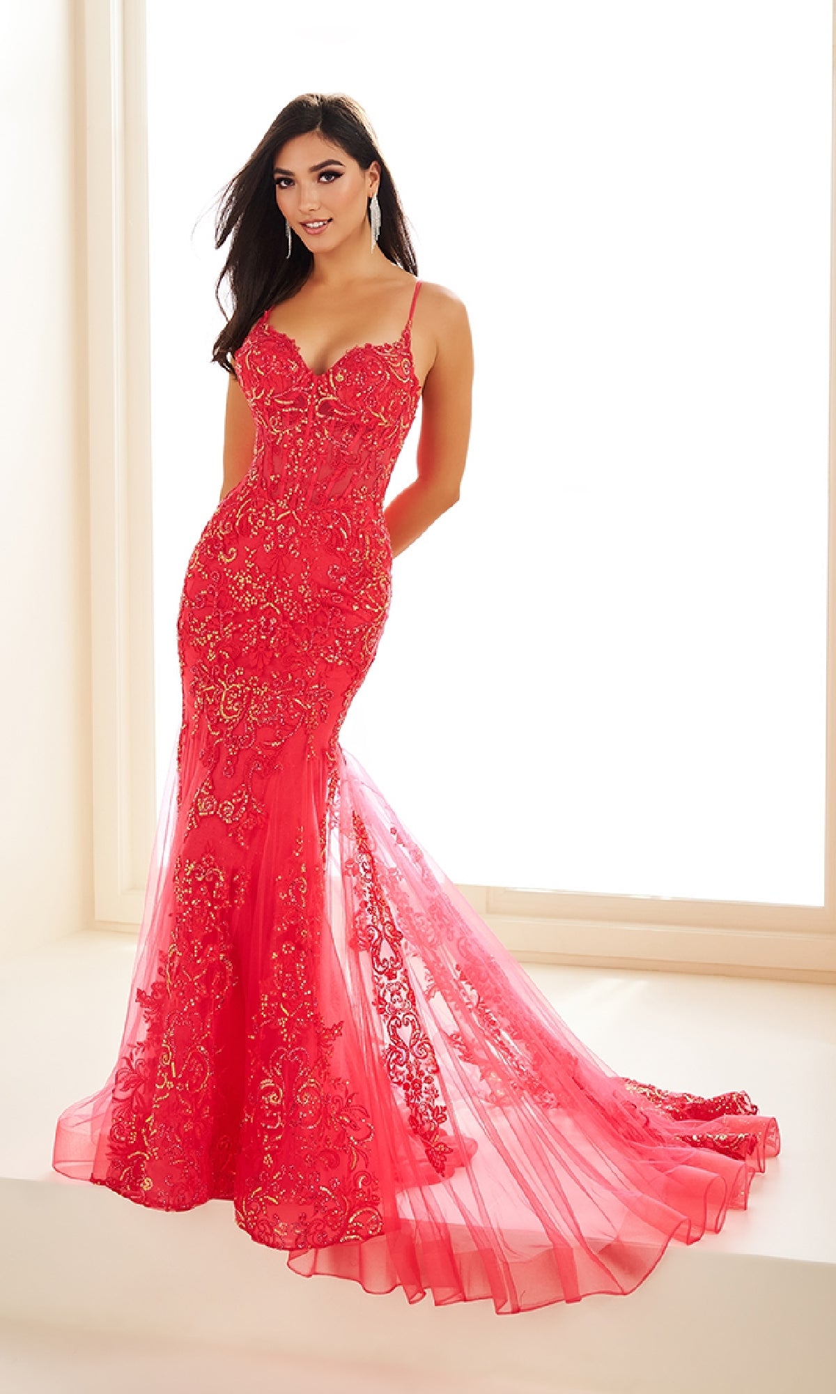 A woman stands indoors wearing the Ellie Wilde EW36069, a fitted, floor-length red glitter tulle mermaid prom dress with gold lace appliqué, thin straps, sheer corset bodice, and flared semi-sheer train.